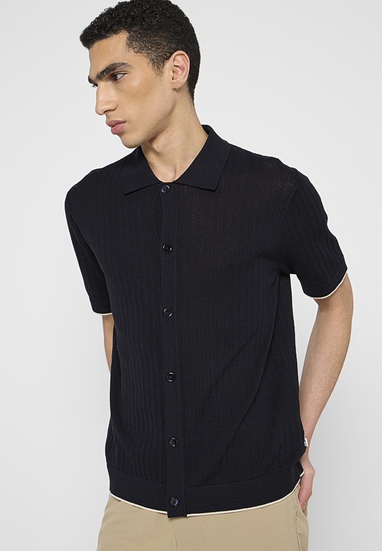 Młody mężczyzna z kolczykiem, ubrany w czarną koszulę z krótkim rękawem i beżowe spodnie, patrzy w bok na jednolitym tle.