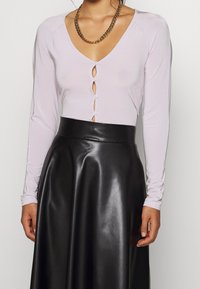 Frau in einem helllila, mit Knöpfen versehenen Oberteil mit Schlüsselloch-Ausschnitten und einem hoch taillierten schwarzen Lederrock, accessoriert mit einer goldenen Kettenhalskette.