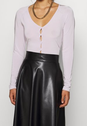 Vrouw in een lichtpaarse blouse met knopen en sleutelgaatjes, gecombineerd met een hoog in de taille zittende zwarte leren rok, accessorized met een gouden ketting.