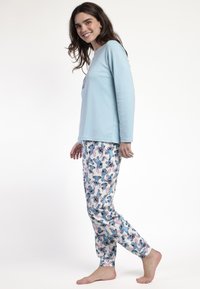 Mujer sonriente con una blusa de manga larga azul claro y pantalones de pijama blancos con estampado de personaje de dibujos animados en azul, de pie descalza contra un fondo blanco.