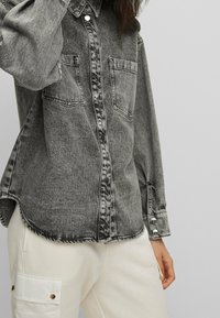 Chemise en denim gris avec deux poches frontales, fermeture par boutons et surpiqûres contrastantes. Le tissu présente un aspect légèrement délavé et texturé.