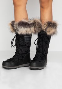 Piernas vistiendo botas de invierno Moon Boot negras con borde de piel y cordones cruzados sobre un suelo blanco y un fondo liso.