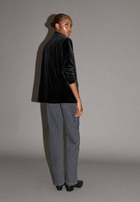 Blazer in velluto nero con maniche lunghe, abbinato a jeans grigi a gamba dritta. Modello mostrato di spalle su uno sfondo grigio.