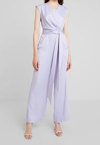 Lila jumpsuit med djup V-ringning, ärmlös design, knytning i midjan och vida byxor. Mjuk textur med draperade fickor.