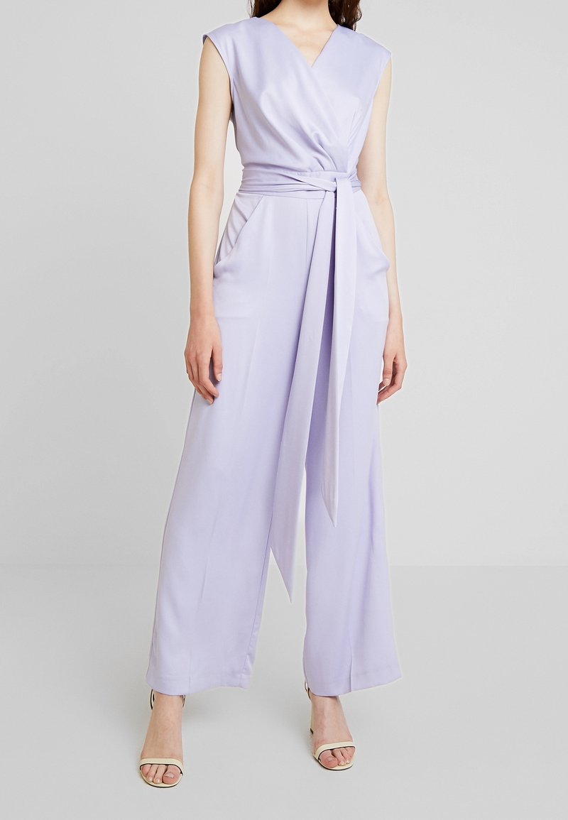 Lila jumpsuit med djup V-ringning, ärmlös design, knytning i midjan och vida byxor. Mjuk textur med draperade fickor.