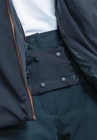 Marineblaues Jacket mit einem Messingreißverschluss und einer versteckten Druckknopfverschluss-Tasche. Glatte Textur mit einem kontrastierenden Innenfutter.