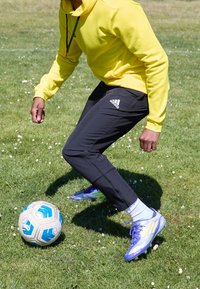 Sweat à capuche jaune vif, pantalon de sport noir et chaussures de football bleues et argentées. En train de donner un coup de pied à un ballon de football bleu et blanc sur de l'herbe verte.