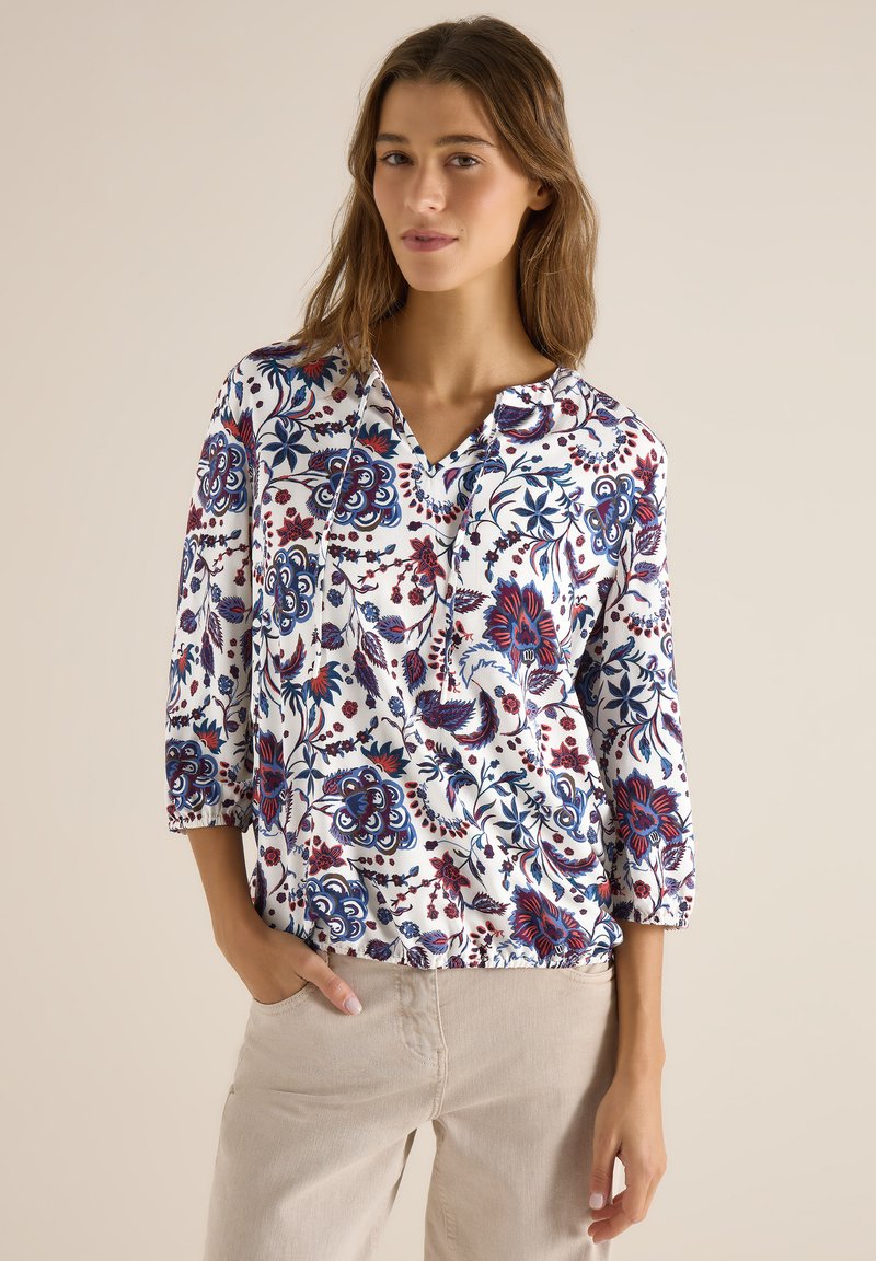 Femme portant un chemisier blanc à motifs floraux bleus et rouges et un pantalon beige, posant avec une main dans la poche sur un fond uni.