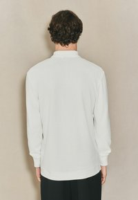 Balta, ilga rankovė polo marškinėliai su grublėta tekstūra, apykakle ir rankogaliais. Turi tiesų pašalą, tinkama neformaliam dėvėjimui.