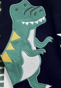 Tengerészeti kötött anyag, zöld hímzett dinoszaurusz mintával, fehér hasával, sárga tüskéivel és részletes arcvonásaival.