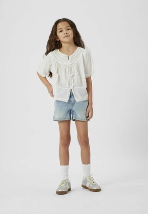 Blouse blanche à manches bouffantes, avec des détails en dentelle et un lien à l'avant, associée à un short en denim bleu clair et des baskets argentées avec des chaussettes blanches.