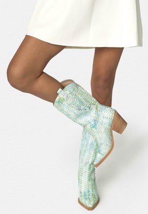 Piernas con botas hasta mitad de la pantorrilla con estampado iridiscente de serpiente en tonos azul verdosos y tacones de madera apilados, combinadas con una falda blanca.