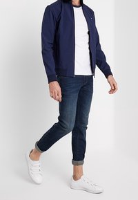 Marinblå bomberjacka över en vit t-shirt, kombinerad med mörkblå jeans och vita sneakers. Outfiten har en avslappnad, modern design.