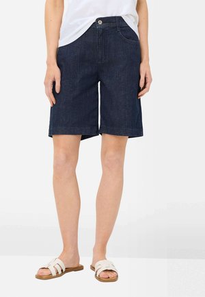 Person iført mørkeblå denimshorts, hvid t-shirt og hvide flade slidesandaler, stående mod en hvid baggrund.