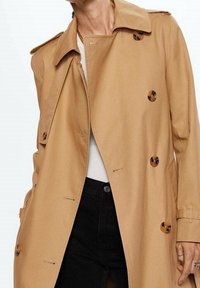 Beżowy płaszcz trench z gładkiego materiału, z podwójnym zapięciem, dużymi guzikami i paskiem w talii. Otwarty kołnierz i kieszenie boczne.