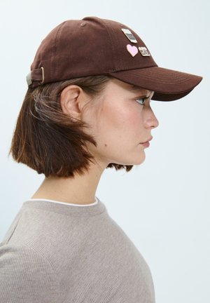 Cappello marrone in twill di cotone, con vari distintivi sulla parte anteriore, tra cui un cuore e un badge, dotato di visiera curva e cinturino regolabile.