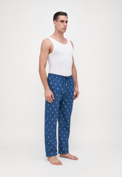 Pantalon de pyjama bleu avec des motifs de chevaux blancs, taille élastique et cordon de serrage, associé à un débardeur blanc, porté pieds nus sur un fond neutre.