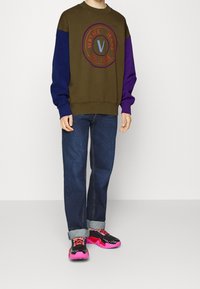 Olivgrön och lila färgblockerad sweatshirt med en rund logotyp. Kombinerad med blå denimjeans och svart-rosa sneakers.