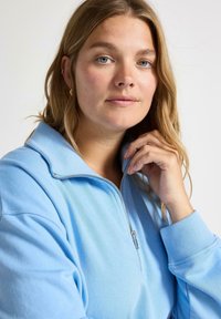 Jonge vrouw met lichtbruin haar, gekleed in een lichtblauwe sweatshirt met rits, die de kraag aanraakt, tegen een plain witte achtergrond.