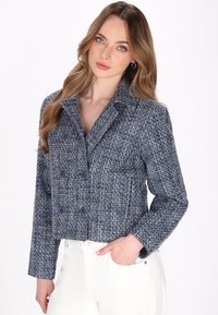 Przycięta granatowa marynarka z tkaniny tweed, z teksturowanym wzorem, z wykładanym kołnierzem, podwójnym zapięciem i dwoma przednimi kieszeniami. Widoczne są dwa niebieskie guziki.