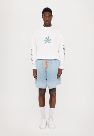 Weißes Langarmshirt mit blauer Grafik auf der Brust und bedrucktem Text auf dem Ärmel, kombiniert mit hellblauen Shorts und weißen Sneakers.