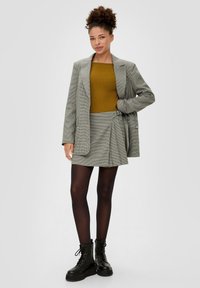 Grau kariertes Blazer über einem senfgelben gerippten Oberteil, kombiniert mit einem grauen Karorock. Das Model trägt schwarze Strumpfhosen und klobige schwarze Combat Boots.
