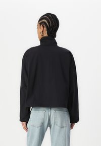 Giacca nera cropped con colletto alto, maniche lunghe e tessuto morbido, abbinata a jeans di denim azzurri chiari.