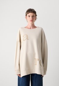 Beige oversized sweatshirt met een brede halslijn en lange mouwen, met subtiele lichtbruine opdruk aan de voorkant. Blauwe spijkerbroek zichtbaar.