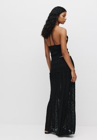 Haut court noir à fines bretelles, associé à une longue jupe maxi noire transparente avec des textures superposées et des détails plissés.