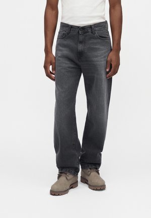 Homme portant un jean noir délavé droit, des bottes beiges à lacets et un t-shirt blanc, debout devant un fond blanc uni.