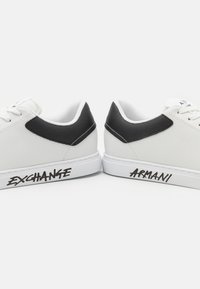 Hvite sneakers med svarte teksturerte detaljer, med snøring og merking "EXCHANGE" og "ARMANI" på sidene.