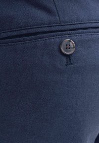 Primo piano di pantaloni in tessuto blu scuro che mostrano una tasca posteriore con bottone, cuciture visibili e trama strutturata.