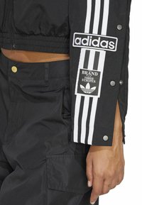 Jachetă neagră scurtă cu trei dungi albe, patch cu logo-ul Adidas și capse pe mâneci. Material ușor, cu textură netedă.