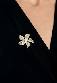 Gold-tone floral brooch with a central pearl, featuring rhinestone accents and leaf-patterned petals. Attached to a black fabric garment.