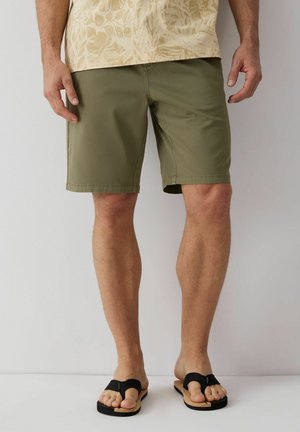 Mężczyzna ubrany w oliwkowo-zielone szorty i czarne sandały, stojący na jednolitym białym tle.