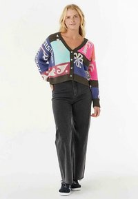 Cardigan court multicolore avec motifs géométriques ; fabriqué en maille, avec un col en V profond et des fermetures à boutons, porté avec un jean foncé.