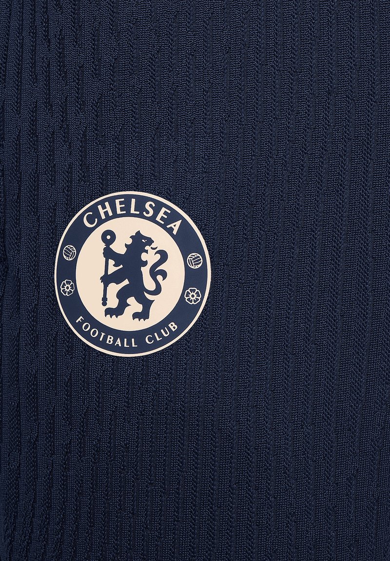 Suéter de color azul marino con textura acanalada que presenta un logo circular del Chelsea Football Club en beige claro, que representa un león y iconos deportivos.