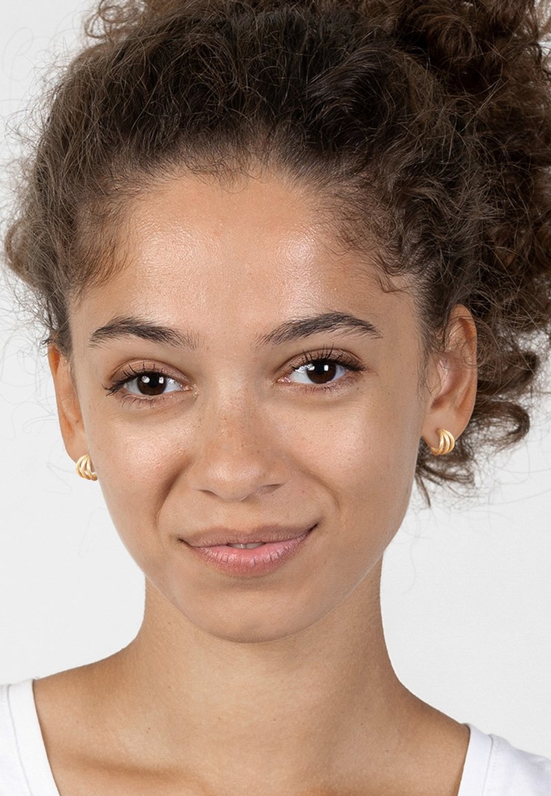 Guldörhängen i form av hoops med en tvinnad design, som har en slät yta och glänsande finish. Personen har lockigt hår och en ljus skjorta.