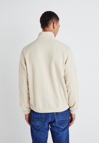 Beige fleece jacket with a high collar, long sleeves, and elastic cuffs, paired with blue denim jeans and visible back pockets.