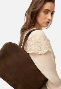 Brown suede handbag with a rectangular shape, featuring a smooth texture and two sturdy top handles, worn on the shoulder against a light blouse.