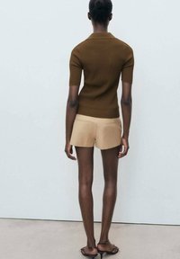 Hnědý žebrovaný top s krátkým rukávem v kombinaci s béžovými šortkami, které prezentují přiléhavý design a minimalistický styl. Model se otáčí zády k divákovi.