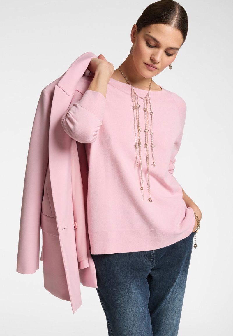 Maglione rosa chiaro con scollatura rotonda e maniche lunghe, adornato con una collana a catena a strati. Indossato con un blazer rosa abbinato e jeans scuri.