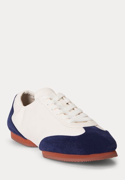 Baskets blanches avec accents en daim bleu marine sur l\'avant et le talon, lacets blancs et semelle en caoutchouc marron, présentées légèrement en angle sur fond blanc.