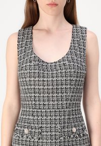 Mustavalkoinen tweed-mekko, jossa on pyöreä pääntie, hihaton muotoilu, teksturoitu kangas ja kaksi taskua, joissa on pyöreitä koristenappeja.