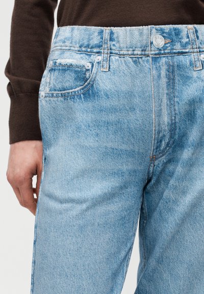 Ανοιχτό μπλε τζιν παντελόνι με μπροστινές τσέπες και κλείσιμο με κουμπί, συνδυασμένο με μακρυμάνικη μπλούζα σε σκούρο καφέ χρώμα, με το χέρι ακουμπισμένο στο πλευρό.