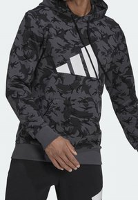 Homme portant un sweat à capuche camouflage noir et gris avec le logo Adidas blanc et un pantalon noir avec des détails blancs, debout devant un fond neutre.