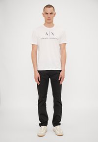 Hvit t-skjorte med rund hals og marineblå trykt logo "A|X ARMANI EXCHANGE," sammen med svarte bukser og hvite sneakers med lysebrune såler.