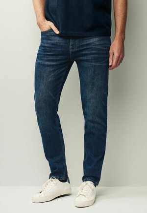 Mężczyzna ubrany w ciemnoniebieskie obcisłe dżinsy, białe trampki i czarną koszulę, stojący z jedną ręką w kieszeni na jednolitym tle.