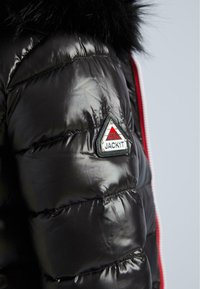 Detailní záběr na černou lesklou bundu s kapucí, na rukávu je trojúhelníková nášivka s logem "JACKIT" a černý kožich na límci.