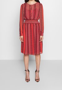 Robe rouge à motifs avec des manches longues, taille cintrée et jupe arrivant au genou. Présente un mélange de motifs linéaires et ovales sur un tissu transparent.